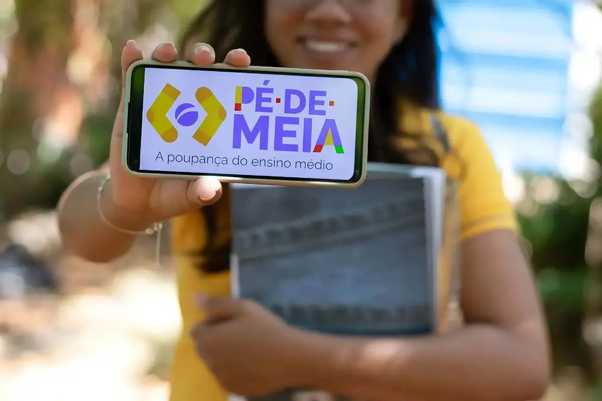 Estudante sorridente do ensino médio público segurando um celular com o aplicativo Jornada do Estudante, representando o programa Pé-de-Meia 2025 no Brasil.