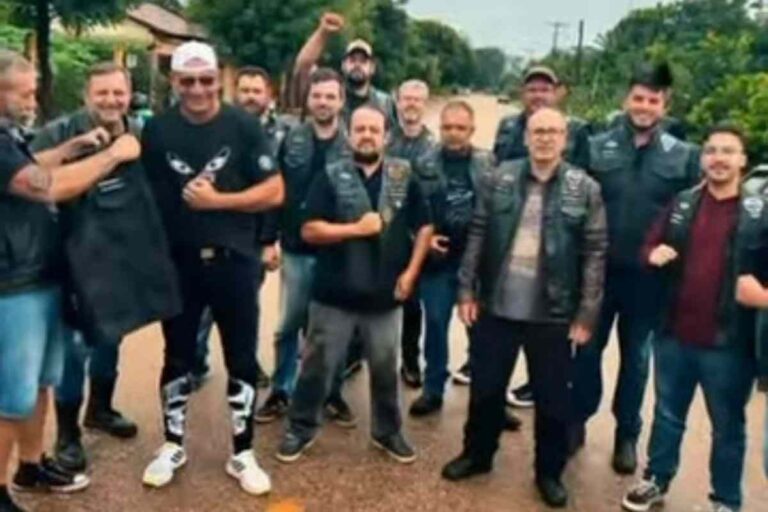 Grupo de motociclistas do Moto Clube Caveiras do Cerrado celebrando a 1ª Feijoada em Rondonópolis, com motos ao fundo e uma mesa repleta de feijoada.