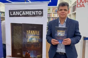 Autor Misael Lopes no lançamento do livro Hannah e os Vampiros da Floresta em Curitiba, segurando a obra em uma livraria com banner de divulgação ao fundo.