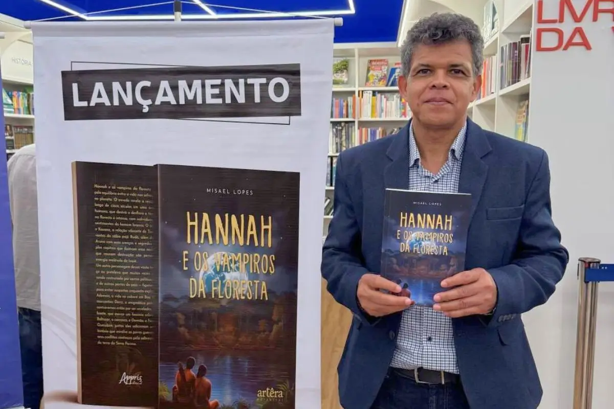 Autor Misael Lopes no lançamento do livro Hannah e os Vampiros da Floresta em Curitiba, segurando a obra em uma livraria com banner de divulgação ao fundo.