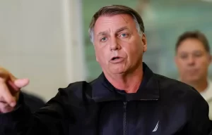 Jair Bolsonaro de terno escuro olha para baixo enquanto coloca tornozeleira eletrônica no tornozelo