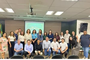 Um grupo de pessoas posa para uma foto oficial em um auditório. Elas estão organizadas em duas fileiras: uma em pé ao fundo e outra agachada ou sentada à frente. Ao centro, há uma tela de projeção com um slide verde que diz: "Agora é hora de ser reconhecida". O ambiente tem iluminação clara de escritório, paredes em tons de cinza e marrom, e uma bandeira do Brasil parcialmente visível à esquerda.