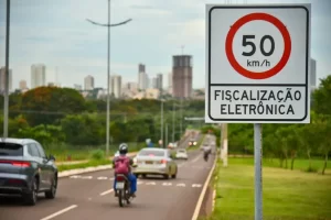 Placa de trânsito em primeiro plano indicando limite de velocidade de 50 km/h e a inscrição "FISCALIZAÇÃO ELETRÔNICA". Ao fundo, uma avenida com carros e motocicletas em movimento, cercada por árvores e prédios ao horizonte sob um céu nublado.