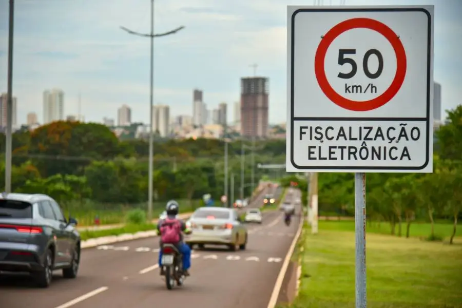 Placa de trânsito em primeiro plano indicando limite de velocidade de 50 km/h e a inscrição "FISCALIZAÇÃO ELETRÔNICA". Ao fundo, uma avenida com carros e motocicletas em movimento, cercada por árvores e prédios ao horizonte sob um céu nublado.