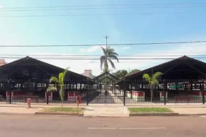 Fotografia da fachada da Feira da Vila Aurora durante o dia. A estrutura é composta por dois grandes pavilhões simétricos com telhados escuros e bases pintadas em vermelho e branco. No centro, um corredor de entrada com portão de ferro preto e palmeiras decorativas nas calçadas laterais. O céu está limpo e azul ao fundo.