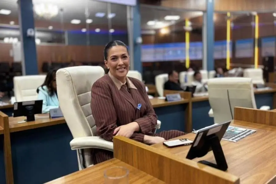 Fotografia da vereadora Luciana Horta sentada em sua mesa no plenário da Câmara Municipal. Ela sorri para a câmera, transmitindo uma postura de liderança e presença no ambiente legislativo.