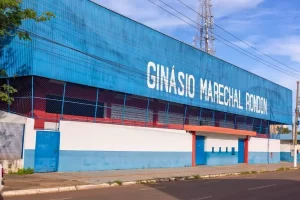 Fachada do Ginásio Marechal Rondon. O edifício tem uma estrutura superior em metal azul com o nome do ginásio escrito em letras brancas grandes. A base é um muro pintado de branco e azul, com portões metálicos. Ao fundo, vê-se uma torre de comunicações e o céu claro.