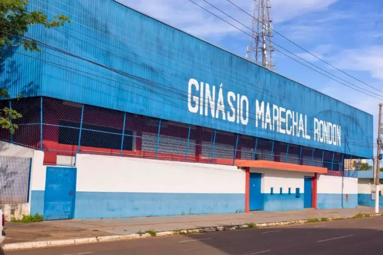 Fachada do Ginásio Marechal Rondon. O edifício tem uma estrutura superior em metal azul com o nome do ginásio escrito em letras brancas grandes. A base é um muro pintado de branco e azul, com portões metálicos. Ao fundo, vê-se uma torre de comunicações e o céu claro.