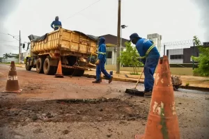 Trabalhadores da prefeitura realizam manutenção asfáltica em uma rua. Um caminhão caçamba amarelo está parado ao lado de um grande buraco no asfalto. Três homens com uniformes azuis e faixas refletivas trabalham no local: um varre a área, outro caminha próximo ao caminhão e o terceiro está em cima da caçamba. Cones de sinalização laranjas delimitam o espaço de trabalho sob um céu nublado.