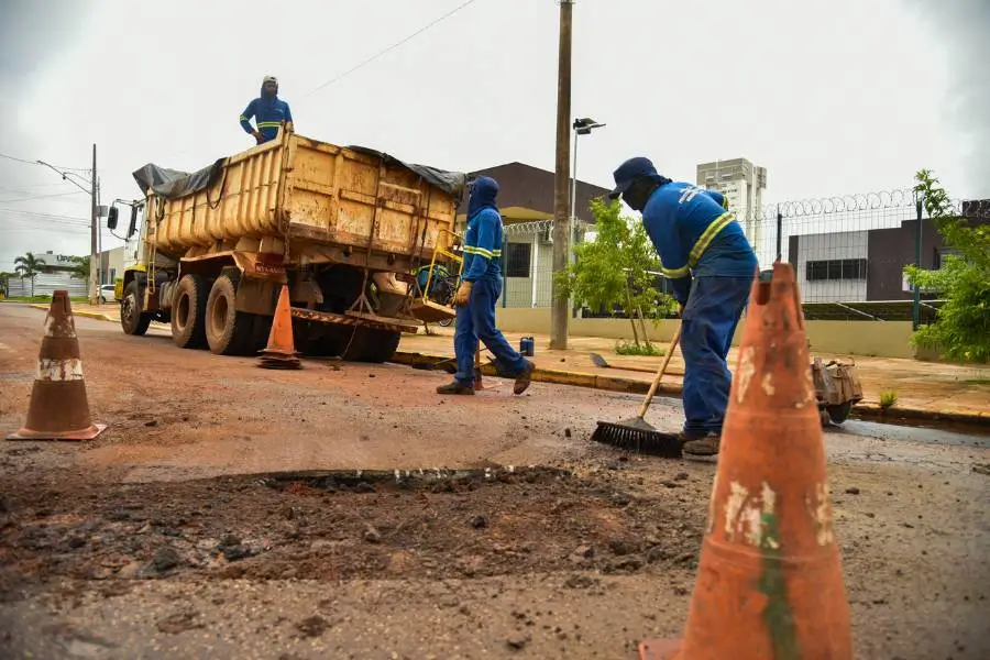 Trabalhadores da prefeitura realizam manutenção asfáltica em uma rua. Um caminhão caçamba amarelo está parado ao lado de um grande buraco no asfalto. Três homens com uniformes azuis e faixas refletivas trabalham no local: um varre a área, outro caminha próximo ao caminhão e o terceiro está em cima da caçamba. Cones de sinalização laranjas delimitam o espaço de trabalho sob um céu nublado.