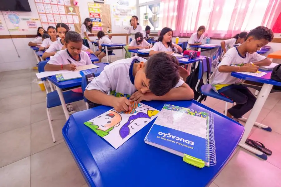 A imagem mostra uma sala de aula com alunos do ensino fundamental sentados em carteiras azuis, concentrados em uma atividade de pintura e desenho. Um dos estudantes aparece em destaque, colorindo uma ilustração, enquanto os demais realizam tarefas em seus cadernos. O ambiente possui cartazes educativos nas paredes e janelas com cortinas ao fundo.