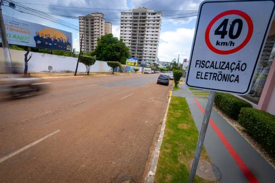 Fotografia colorida em plano médio de uma calçada em Rondonópolis. À direita, em destaque, há uma placa branca retangular com a inscrição '40 km/h' dentro de um círculo vermelho e, abaixo, o texto 'FISCALIZAÇÃO ELETRÔNICA'. A rua ao lado está levemente desfocada, sugerindo movimento, com prédios altos e árvores ao fundo sob um céu nublado.