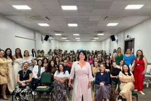 Fotografia de um encontro presencial do grupo Mulheres Empreendedoras em um grande auditório. Em primeiro plano, uma mulher com vestido claro sorri para a câmera, representando a liderança. Atrás dela, várias mulheres de diversas idades estão sentadas em fileiras de cadeiras verdes, preenchendo todo o ambiente. Algumas mulheres também estão em pé nas laterais. A iluminação é clara e o clima é de união e engajamento coletivo.