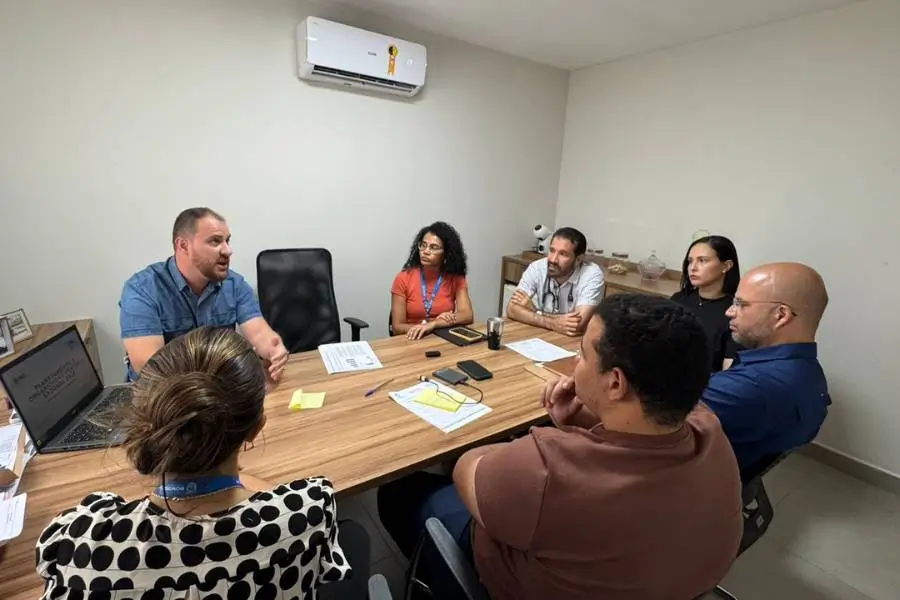 Grupo de seis pessoas participa de reunião em sala de escritório, sentados ao redor de uma mesa com documentos e notebook.