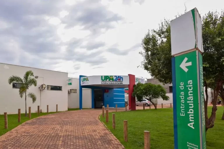 Fachada de uma Unidade de Pronto Atendimento (UPA 24h). Em primeiro plano à direita, um totem verde indica a entrada de ambulâncias com uma seta. Ao fundo, o prédio térreo com detalhes em azul e vermelho, cercado por gramado