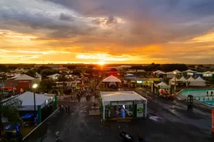Aprosoja MT intensifica presença em feiras para fortalecer diálogo com produtor rural 1 Foto aérea panorâmica de uma grande feira agropecuária ao entardecer. Em primeiro plano, vê-se a estrutura de estandes da Aprosoja MT, organizados para receber produtores. Ao fundo, o parque de exposições conta com diversas tendas, máquinas agrícolas e grande circulação de pessoas sob a luz dourada do pôr do sol.