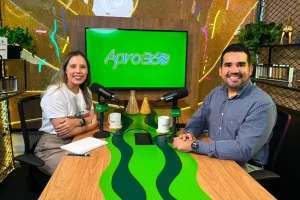Foto em plano médio de um homem e uma mulher sentados em uma bancada de estúdio de podcast. À esquerda, uma mulher de cabelos longos e castanhos veste uma camiseta branca e calça clara. À direita, um homem de barba, vestindo uma camisa social azul de mangas compridas, sorri para a câmera. Entre eles, há uma bancada de madeira com um detalhe em verde ondulado, onde estão dois microfones Shure com o logotipo "Apro360" e duas canecas brancas. Ao fundo, uma tela de TV exibe o logotipo verde e branco "Apro360", com decoração de grãos em frascos de vidro e iluminação neon.