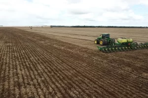 Fotografia aérea de uma vasta área agrícola sob um céu nublado. À direita, um trator verde acoplado a uma plantadeira amarela e preta opera em um terreno que apresenta faixas de terra escura e úmida intercaladas com restos de palhada seca. Ao fundo, a linha do horizonte revela a imensidão da propriedade rural.