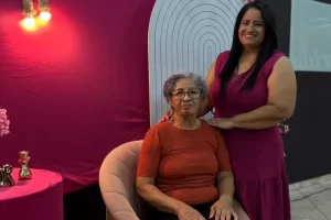 Foto em plano médio de duas mulheres em um ambiente festivo com decoração em tons de rosa. À esquerda, uma senhora de cabelos curtos grisalhos e óculos, vestindo blusa terracota, está sentada em uma poltrona de veludo rosada. À direita, uma mulher de cabelos longos e lisos, vestindo um vestido rosa-escuro, está em pé ao lado da senhora com as mãos gentilmente apoiadas em seus ombros. Ao fundo, uma parede rosa com iluminação decorativa e uma estrutura em arco.