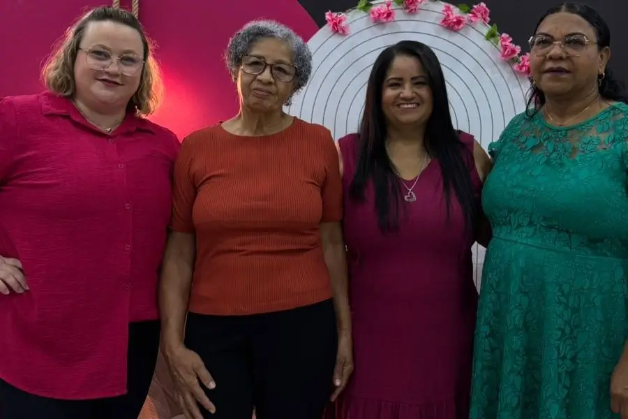 Foto de quatro mulheres posando juntas, lado a lado, em um ambiente interno. A mulher à esquerda usa uma camisa de botão em um tom de rosa vibrante e óculos. A segunda mulher, ao centro-esquerda, tem cabelos grisalhos curtos, usa óculos e uma blusa em tom terracota. A terceira mulher, ao centro-direita, tem cabelos longos e escuros, usa um vestido em um tom de rosa escuro e um colar com pingente. A mulher à direita usa um vestido verde de renda com detalhes florais e óculos. Atrás delas, há uma decoração circular com flores cor-de-rosa.