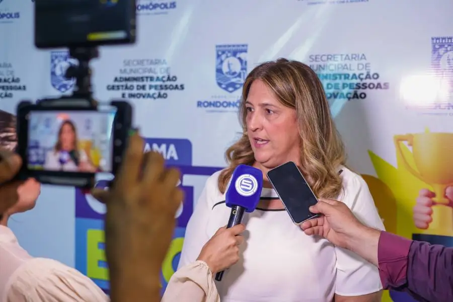 Uma mulher de cabelos loiros e ondulados fala diante de microfones e celulares em uma entrevista. Ela está vestindo uma blusa branca. Ao fundo, vê-se um painel com a logomarca da Secretaria Municipal de Administração, Gestão de Pessoas e Inovação da Prefeitura de Rondonópolis. Em primeiro plano, parte dos braços e das mãos de profissionais de imprensa segurando os equipamentos de gravação.