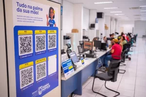 Painel informativo vertical com fundo azul e branco, posicionado no interior de um escritório de atendimento da prefeitura. O painel traz o título "Tudo na palma da sua mão!" e apresenta uma série de QR Codes para a emissão de documentos como IPTU, BCI, BCM e Certidões. Ao fundo, desfocado, vê-se um balcão de atendimento com funcionários trabalhando e alguns cidadãos sentados aguardando atendimento em um ambiente claro e organizado.