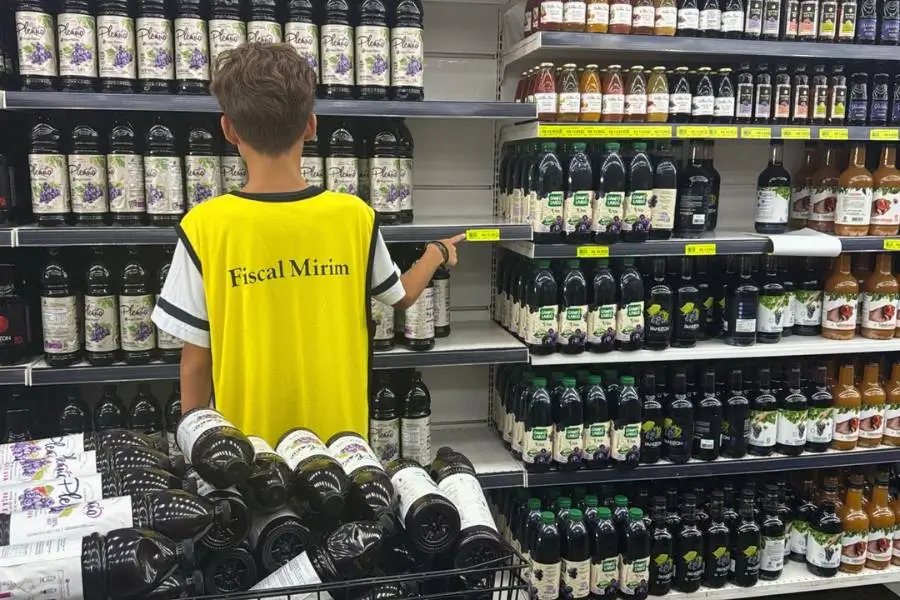 A imagem mostra um menino de costas, usando um colete amarelo com a frase "Fiscal Mirim" escrita em preto. Ele está em um corredor de supermercado e aponta com o dedo indicador para uma etiqueta de preço amarela em uma prateleira repleta de garrafas de suco de uva. Ao lado dele, há um carrinho metálico também cheio de garrafas.