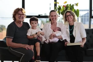 Produtora rural de Tapurah, Inez Catelan Lazarotto constrói uma história de superação no campo 1 Foto em plano médio de três mulheres adultas e duas crianças sentadas em um sofá preto. Da esquerda para a direita: uma mulher mais velha de cabelos curtos e óculos; ao seu lado, um menino pequeno sentado no colo de uma mulher jovem de cabelos castanhos, que também segura um bebê. À direita, uma terceira mulher usa blazer branco, colar de pedras azuis e segura um livro ou pasta. Todas sorriem para a câmera em um ambiente iluminado por luz natural de uma grande janela ao fundo.