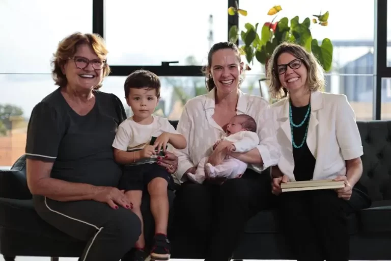 Foto em plano médio de três mulheres adultas e duas crianças sentadas em um sofá preto. Da esquerda para a direita: uma mulher mais velha de cabelos curtos e óculos; ao seu lado, um menino pequeno sentado no colo de uma mulher jovem de cabelos castanhos, que também segura um bebê. À direita, uma terceira mulher usa blazer branco, colar de pedras azuis e segura um livro ou pasta. Todas sorriem para a câmera em um ambiente iluminado por luz natural de uma grande janela ao fundo.