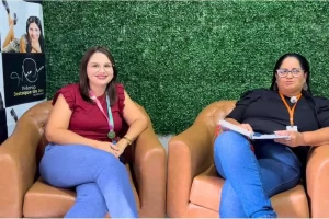 Duas mulheres estão sentadas em poltronas de couro marrom. A mulher à esquerda tem cabelos castanhos lisos, usa óculos e veste uma blusa bordô. A mulher à direita tem cabelos pretos, usa óculos de armação escura e veste uma blusa preta, segurando um caderno. Ao fundo, uma parede de folhagens verdes e um banner à esquerda com a inscrição "Prêmio Destaque do Ano".