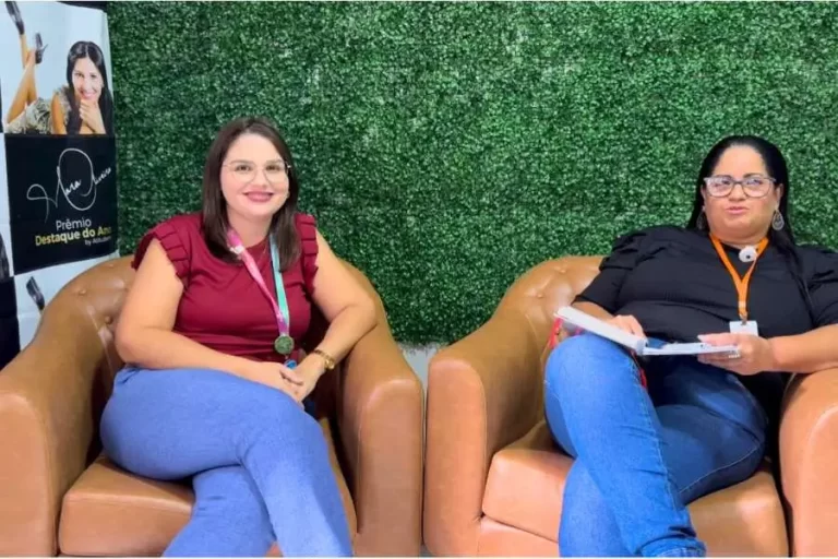 Duas mulheres estão sentadas em poltronas de couro marrom. A mulher à esquerda tem cabelos castanhos lisos, usa óculos e veste uma blusa bordô. A mulher à direita tem cabelos pretos, usa óculos de armação escura e veste uma blusa preta, segurando um caderno. Ao fundo, uma parede de folhagens verdes e um banner à esquerda com a inscrição "Prêmio Destaque do Ano".