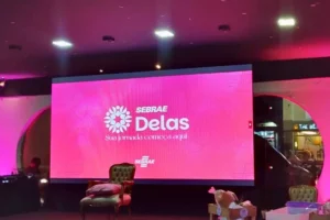 Uma tela de LED grande e vibrante em tons de rosa exibe no centro o logotipo do "SEBRAE Delas" em branco, com uma flor estilizada à esquerda do nome e a frase "Sua jornada começa aqui" logo abaixo. O palco em primeiro plano está vazio, com uma poltrona clássica capitonê de couro verde oliva e uma almofada rosa sobre ela. Ao fundo, um ambiente de evento com iluminação de teto e decorações laterais arqueadas e iluminadas em rosa.