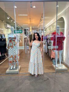 Fotografia colorida da empresária Ana Paula em frente à sua loja, a Manu Modas. Ana Paula tem cabelos pretos ondulados e usa um vestido longo com estampa floral delicada. Ao fundo, o interior da loja é iluminado, com araras de roupas e piso amadeirado. Nas vitrines laterais, manequins exibem vestidos coloridos. Nas portas de vidro, há adesivos escrito 'ABRIR' e uma placa verde de 'ABERTO.