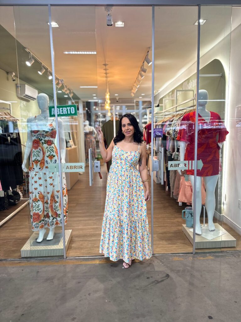 Fotografia colorida da empresária Ana Paula em frente à sua loja, a Manu Modas. Ana Paula tem cabelos pretos ondulados e usa um vestido longo com estampa floral delicada. Ao fundo, o interior da loja é iluminado, com araras de roupas e piso amadeirado. Nas vitrines laterais, manequins exibem vestidos coloridos. Nas portas de vidro, há adesivos escrito 'ABRIR' e uma placa verde de 'ABERTO.