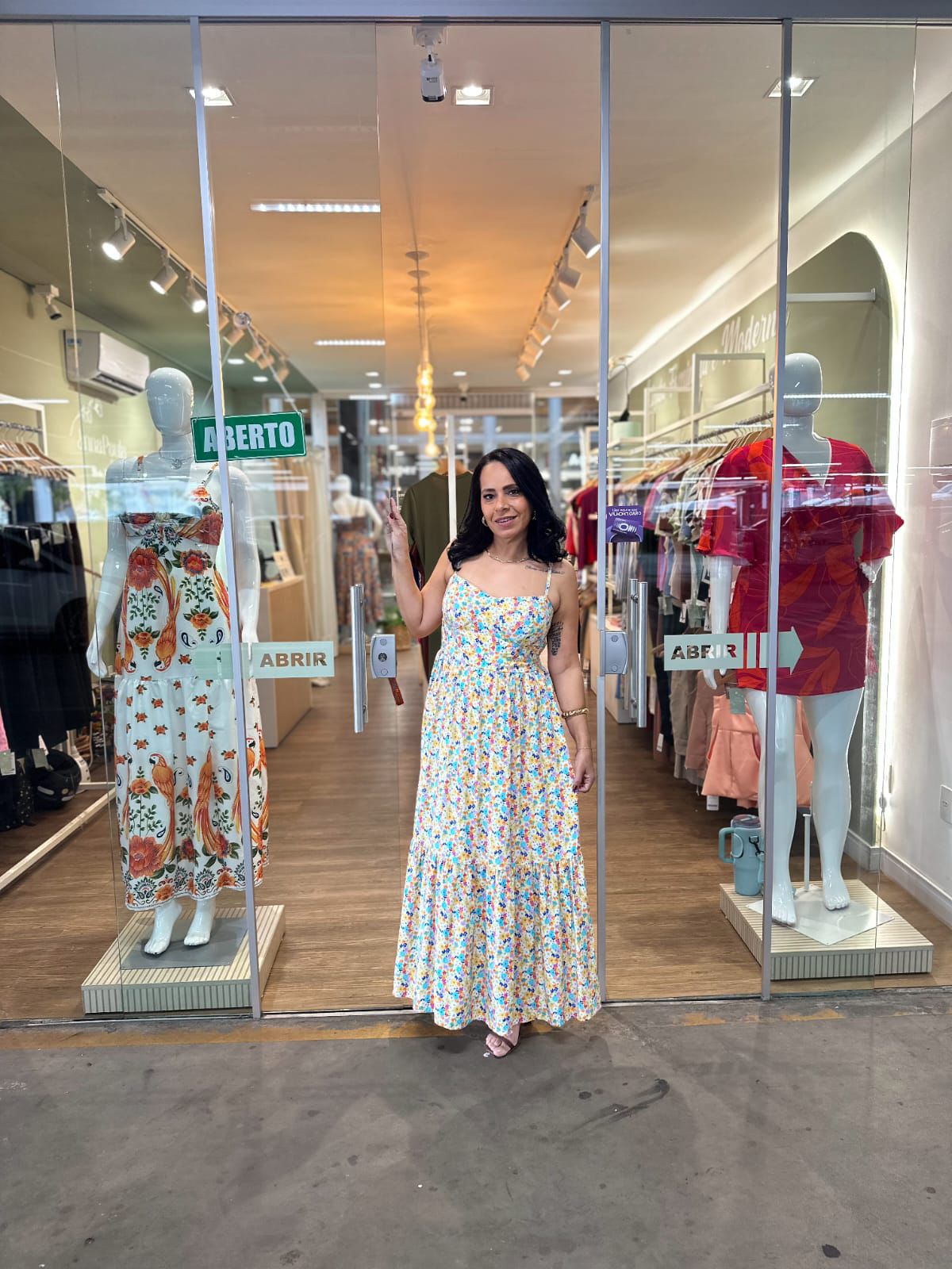 Fotografia colorida da empresária Ana Paula em frente à sua loja, a Manu Modas. Ana Paula tem cabelos pretos ondulados e usa um vestido longo com estampa floral delicada. Ao fundo, o interior da loja é iluminado, com araras de roupas e piso amadeirado. Nas vitrines laterais, manequins exibem vestidos coloridos. Nas portas de vidro, há adesivos escrito 'ABRIR' e uma placa verde de 'ABERTO.