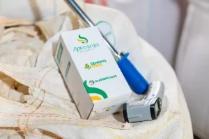 Uma fotografia em close, com profundidade de campo rasa, foca em uma caixa branca de formato trapezoidal sobre um grande saco de ráfia texturizado e cheio de sementes (aparentemente soja). A caixa tem impressos o logotipo verde e amarelo da Aprosoja Mato Grosso, a inscrição verde "SEMENTE FORTE" e, abaixo, a inscrição verde "CLASSIFICADOR". Ao lado da caixa, em pé e ligeiramente atrás, está uma ferramenta de amostragem metálica (concha ou calador) com um cabo de plástico azul brilhante. À direita e em primeiro plano, encontra-se um scanner de mão digital cinza com um pequeno visor preto. O fundo está desfocado e mostra mais sacos de ráfia. A iluminação é de estúdio, suave e uniforme.