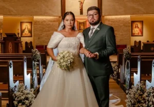 Foto de Mathews e Gabriela de mãos dadas no corredor central da igreja. Ela usa vestido branco ombro a ombro com buquê de rosas; ele veste terno verde-escuro e óculos. Ao fundo, o altar iluminado com imagens sacras e bancos decorados com flores brancas.