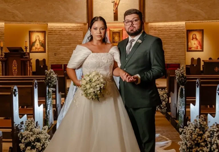 Foto de Mathews e Gabriela de mãos dadas no corredor central da igreja. Ela usa vestido branco ombro a ombro com buquê de rosas; ele veste terno verde-escuro e óculos. Ao fundo, o altar iluminado com imagens sacras e bancos decorados com flores brancas.