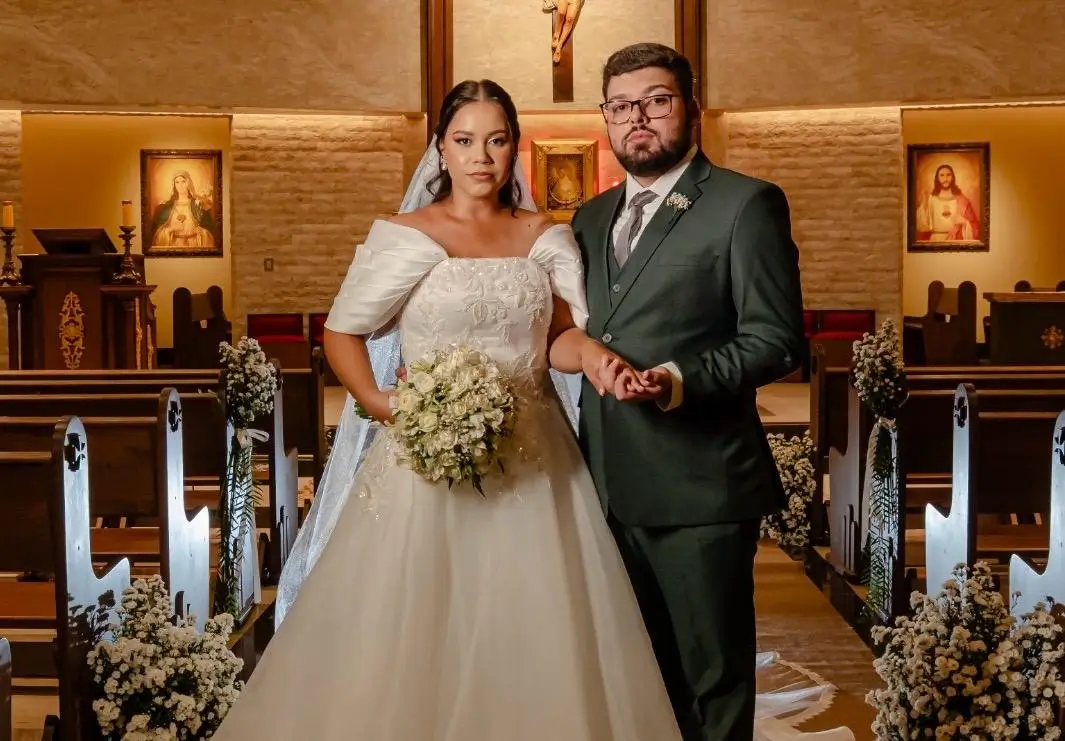 Foto de Mathews e Gabriela de mãos dadas no corredor central da igreja. Ela usa vestido branco ombro a ombro com buquê de rosas; ele veste terno verde-escuro e óculos. Ao fundo, o altar iluminado com imagens sacras e bancos decorados com flores brancas.