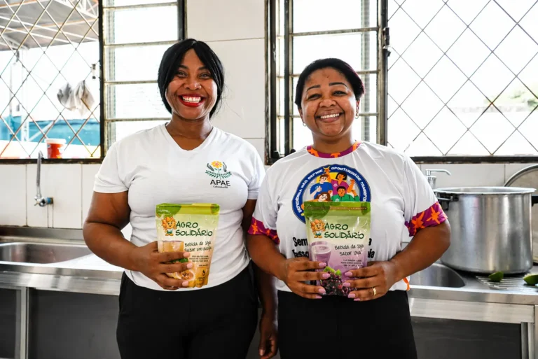Fotografia colorida em plano médio mostra duas mulheres negras, colaboradoras da APAE, sorrindo calorosamente para a câmera. Elas estão lado a lado, em pé, dentro de uma cozinha industrial limpa e bem iluminada por janelas ao fundo. Ambas seguram embalagens do programa "Agrosolidário". A mulher à esquerda veste uma camiseta branca com o logo da APAE de Cáceres; ela segura um pacote amarelo de bebida de soja sabor maracujá. A mulher à direita veste uma camiseta branca com estampas e mangas coloridas; ela segura um pacote roxo de bebida de soja sabor uva. À direita, vê-se uma grande panela de alumínio no fogão.