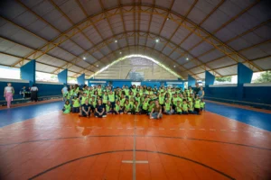 Fotografia em plano aberto mostra um grupo de cerca de 100 alunos e educadores reunidos no centro de uma quadra poliesportiva coberta. Os estudantes vestem coletes amarelos fluorescentes e estão sentados no piso de cor laranja, posicionados para a foto oficial. Ao fundo, a estrutura metálica do telhado e as arquibancadas azuis da escola CMEB completam o cenário. Todos olham para a câmera em um momento de integração.
