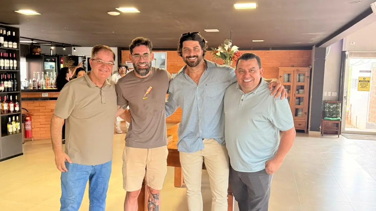 Fotografia colorida em plano médio de quatro homens adultos posicionados lado a lado, sorrindo para a câmera em um ambiente interno iluminado. O homem à esquerda veste óculos e uma camisa polo bege. O segundo homem possui barba, óculos e veste uma camiseta cinza. O terceiro homem, no centro, é alto, possui barba e veste uma camisa social de mangas longas com listras azuis e brancas. O quarto homem, à direita, veste uma camisa polo cinza claro e mantém o braço direito apoiado no ombro do homem ao seu lado. O cenário ao fundo é composto por um balcão de tijolos aparentes, prateleiras com garrafas de vinho e móveis de madeira, sugerindo o interior de um restaurante ou hotel.