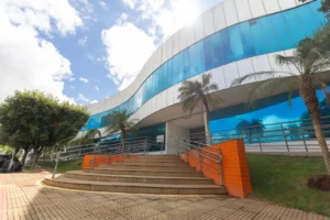 Fotografia em ângulo baixo da fachada da Prefeitura de Rondonópolis. O edifício possui arquitetura moderna e curvilínea, com grandes janelas de vidro espelhado em azul intenso e estrutura branca. À frente, uma escadaria de concreto em leque com corrimãos metálicos leva à entrada. As laterais da escada são revestidas com azulejos laranjas. Palmeiras e árvores verdes decoram o entorno sob um céu ensolarado.