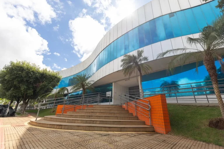 Fotografia em ângulo baixo da fachada da Prefeitura de Rondonópolis. O edifício possui arquitetura moderna e curvilínea, com grandes janelas de vidro espelhado em azul intenso e estrutura branca. À frente, uma escadaria de concreto em leque com corrimãos metálicos leva à entrada. As laterais da escada são revestidas com azulejos laranjas. Palmeiras e árvores verdes decoram o entorno sob um céu ensolarado.