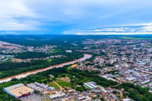 Vista aérea da cidade de Rondonópolis, com um rio de águas barrentas atravessando áreas urbanas e regiões verdes, cercado por bairros, prédios e vegetação.