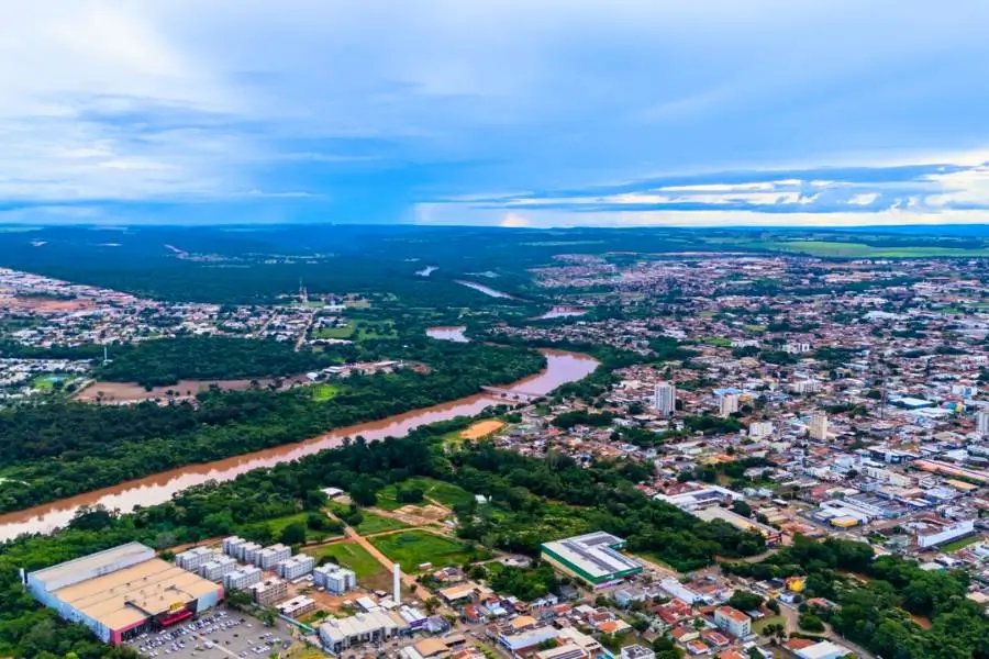 Vista aérea da cidade de Rondonópolis, com um rio de águas barrentas atravessando áreas urbanas e regiões verdes, cercado por bairros, prédios e vegetação.