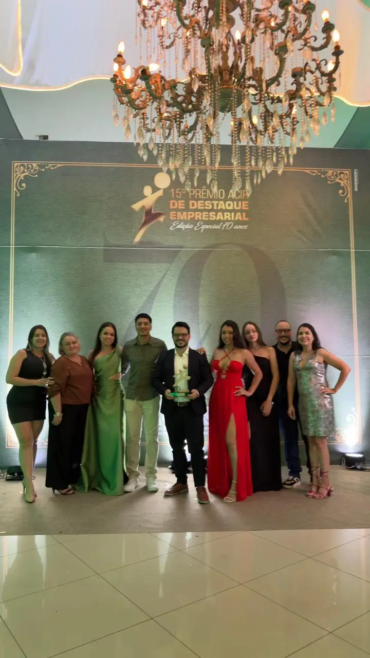 Fotografia colorida vertical em ambiente festivo. No centro da imagem, um grupo de nove pessoas está posicionado à frente de um painel verde escuro com detalhes dourados, onde se lê "15º Prêmio ACIR de Destaque Empresarial - Edição Especial 70 anos". Ao centro, um homem de barba e óculos, vestindo terno azul escuro e camisa branca, segura um troféu dourado. À sua esquerda e direita, homens e mulheres vestem trajes de gala, incluindo vestidos nas cores verde, vermelho, preto e prata brilhante. Acima do grupo, destaca-se um grande e luxuoso lustre de cristal com luzes quentes acesas. O chão é de piso claro e brilhante, refletindo a iluminação do local.