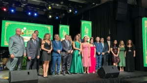 Fotografia colorida em plano médio de um grupo de aproximadamente 18 pessoas, entre homens e mulheres, posicionadas lado a lado em um palco. Os homens vestem ternos e as mulheres usam vestidos de gala em cores variadas, como verde, rosa, vermelho e preto. Ao fundo, três grandes painéis de LED exibem a identidade visual do evento na cor verde com detalhes em dourado, onde se lê "15º Prêmio Destaque Empresarial - Edição Especial 70 anos". O palco tem piso escuro e conta com equipamentos de som (monitores pretos) à frente do grupo e iluminação cênica no teto com refletores coloridos em tons de azul e amarelo. O ambiente é formal e festivo.