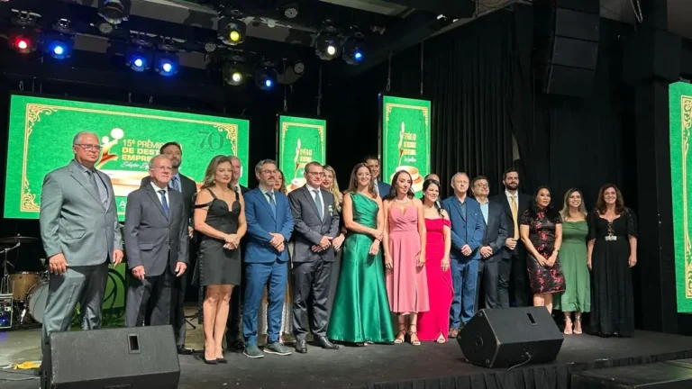 Fotografia colorida em plano médio de um grupo de aproximadamente 18 pessoas, entre homens e mulheres, posicionadas lado a lado em um palco. Os homens vestem ternos e as mulheres usam vestidos de gala em cores variadas, como verde, rosa, vermelho e preto. Ao fundo, três grandes painéis de LED exibem a identidade visual do evento na cor verde com detalhes em dourado, onde se lê "15º Prêmio Destaque Empresarial - Edição Especial 70 anos". O palco tem piso escuro e conta com equipamentos de som (monitores pretos) à frente do grupo e iluminação cênica no teto com refletores coloridos em tons de azul e amarelo. O ambiente é formal e festivo.