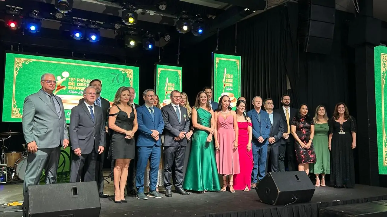 Fotografia colorida em plano médio de um grupo de aproximadamente 18 pessoas, entre homens e mulheres, posicionadas lado a lado em um palco. Os homens vestem ternos e as mulheres usam vestidos de gala em cores variadas, como verde, rosa, vermelho e preto. Ao fundo, três grandes painéis de LED exibem a identidade visual do evento na cor verde com detalhes em dourado, onde se lê "15º Prêmio Destaque Empresarial - Edição Especial 70 anos". O palco tem piso escuro e conta com equipamentos de som (monitores pretos) à frente do grupo e iluminação cênica no teto com refletores coloridos em tons de azul e amarelo. O ambiente é formal e festivo.
